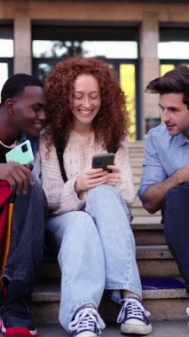 Dikey. Bir grup genç bir okul binasının merdivenlerinde cep telefonlarına bakarken cep telefonu paylaşımı gülümsemelerini kullanarak oturuyorlar. Üniversitede boş bir anın tadını çıkarıyor gibiler.