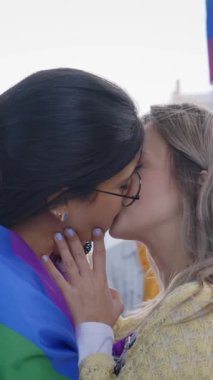Dikey. İki genç lezbiyen kadın LGBT etkinliğinde gökkuşağı bayrağı taşırken öpüşüyorlar. Onların jestleri aynı cinsiyetten insanlar arasındaki sevgi ve eşitliği temsil ediyor..
