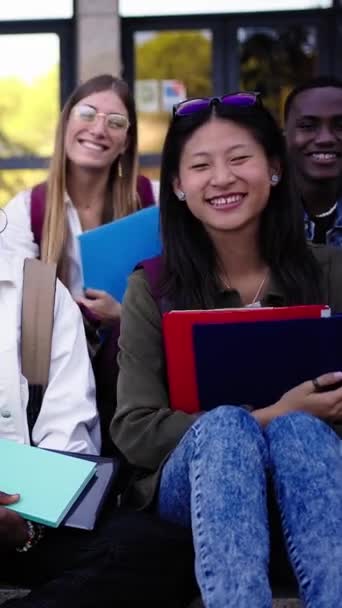 垂直的 多种族的学生坐在楼梯上微笑着 手里拿着文件夹 看着学校外的相机 校园内多文化年轻人同班同学的快乐生活理念 — 图库视频影像