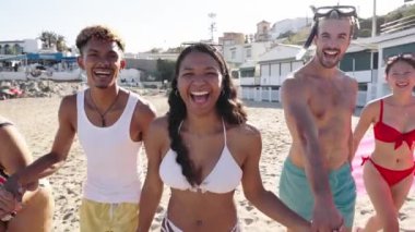 Bir grup heyecanlı arkadaş mayo giymiş bir şehir plajında kumda koşuyor. Genç çok ırklı insanlar yaz tatillerinde plajda eğleniyorlar.