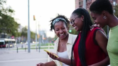 Neşeli Afrikalı kadın arkadaşlar gülen cep telefonuyla karşıdan karşıya geçiyorlar. Genç siyahi kadınlar açık havada cep telefonlarını kullanarak şehirde dolaşmaktan hoşlanırlar. Pozitif sosyal ilişkiler nesli z 