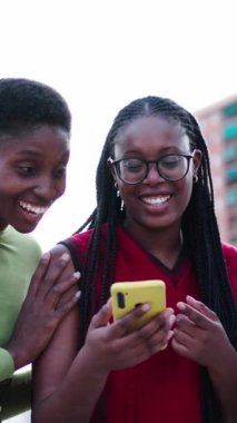 Dikey üç güzel Afro-Amerikan kadını cep telefonundan komik videolar izlerken mutlu bir şekilde gülüyor. Genç siyahi bayan arkadaşlar birlikte boş günün tadını çıkarıyorlar. Hücreleri dışarıda neşeli görünüyor.