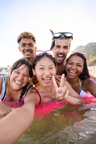 Vertical selfie time. Group of cheerful multiracial friends in swim wear and diving goggles, caps, inflatable mats, are posing for a photo, that Asian lady is taking, beaming smiles, sunshine
