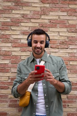 Tuğla duvara yaslanarak gezen yakışıklı bir adamın dikey kopyalama uzayı. Genç, beyaz bir erkek dışarıda cep telefonuna bakıp gülümsemekten zevk alıyor. İnsanlar teknolojiye bağımlı. 