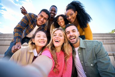 Bir grup çok ırklı arkadaş dışarıda akıllı cep telefonlarıyla selfie çekiyorlar. Mutlu, genç, çok ırklı insanlar kameraya bakarak gülümsüyor. Üniversite öğrencileri üniversite kampüsünde eğleniyor.