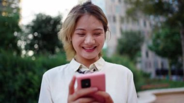 Şehir parkında cep telefonuyla gülümseyen neşeli Çinli genç sarışın kadın. Sosyal medya uygulamasından keyif alan Asyalı güzel bir kız nesli. Dışarıdaki kırmızı cihazlı insanlar ağ bağımlısı oldular 