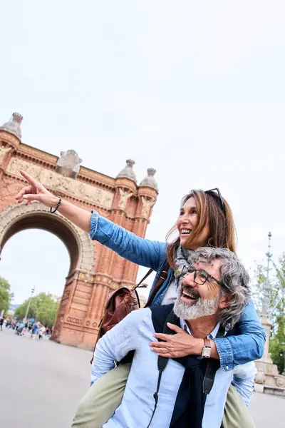 Vertical. Cheerful mature tourist attractive couple pointing at something on city. Man with grey hair gives piggyback ride to smiling woman. Happy adult people in love enjoying sightseeing on vacation