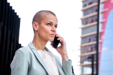 Elektrikli orta yaşlı, tıraşlı bir iş kadını fotokopi alanı için binaların odaksız arka planıyla telefonda konuşuyor. Takım elbiseli çekici kadın profesyoneller iş yerinden ciddi bir telefon görüşmesi yapıyorlar.