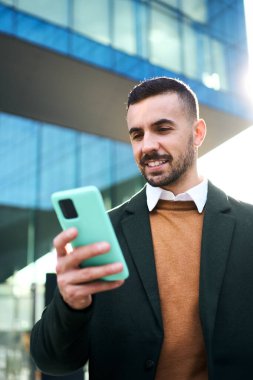 Ofis binasının önünde resmi takım elbiseli genç bir beyaz adamın cep telefonunu kullanıp ona baktığı dikey bir video. İşteki ilk günü için mutlu ve kendinden emin görünüyor.
