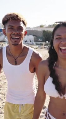 Bir grup heyecanlı arkadaşın dikey fotoğrafı. Bir şehir plajında mayo giyerek kumda koşuyorlar. Genç çok ırklı insanlar yaz tatillerinde plajda eğleniyorlar.