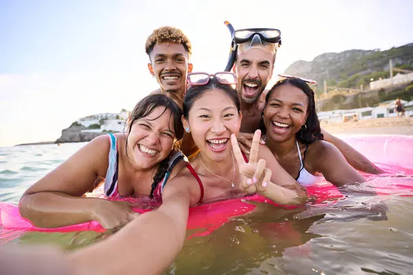Selfie time. Group of cheerful multiracial friends in swim wear and diving goggles, caps, inflatable mats, are posing for a photo, that Asian lady is taking, beaming smiles, sunshine