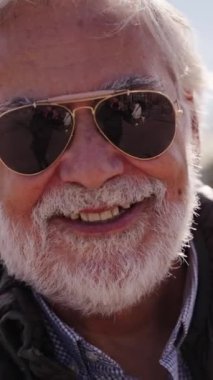 Dikey. Beyaz saçlı, sakallı, mutlu ve gülümseyen beyaz turist portresi güneşli bir kış gününde güneş gözlüğü takarak tatillerinin tadını çıkarıyor.