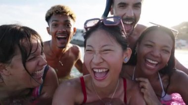 Selfie zamanı. Yüzme kıyafetleri, dalış gözlükleri, şapkalar, şişme paspaslar takan bir grup neşeli çok ırklı arkadaş fotoğraf için poz veriyor. Asyalı kadın gülümsüyor, gün ışığı alıyor.