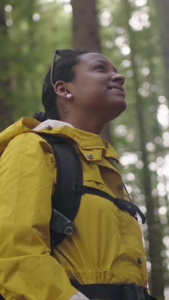 一个身披背包的拉美裔年轻女子的垂直低矮的角度微笑着 在森林中央的大树下欣赏着这一刻 慢动作中的幸福与自由的概念 — 图库视频影像