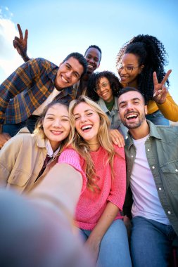 Dikey. Bir grup çok ırklı arkadaş dışarıda akıllı cep telefonlarıyla selfie çekiyorlar. Kameraya bakarak gülümseyen çok ırklı genç insanlar. Üniversite öğrencileri üniversite kampüsünde eğleniyor.