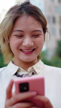 Şehir parkında cep telefonuyla gülümseyen dikey neşeli Çinli genç sarışın kadın. Sosyal medya uygulamasından keyif alan Asyalı güzel bir kadın. Dışarıdaki kırmızı cihazlı insanlar ağ bağımlısı oldular 