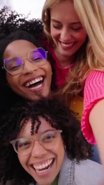 Dikey. Bir grup çok ırklı arkadaş dışarıda akıllı cep telefonlarıyla selfie çekiyorlar. Kameraya bakarak gülümseyen çok ırklı genç insanlar. Üniversite öğrencileri üniversite kampüsünde eğleniyor.