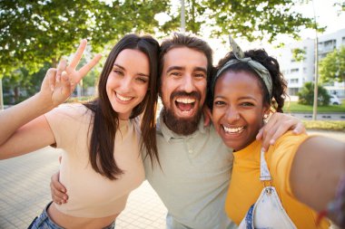 Selfie üç heyecanlı çok ırklı neşeli genç arkadaş dışarıda kucaklaşıyorlar. Kadınlar ve erkekler gülümseyen kameraya bakarak ve yaz tatilinin tadını çıkararak barış sembolü eller yaparak eğleniyorlar. Sunny Park 'taki Gen Z