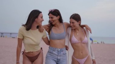 Üç beyaz arkadaş bikinili olarak plajda tatilin tadını çıkarıyorlar. Güneşli bir günde kumsalda yürüyüp gülerken mayo giyip sahilde kaygısız bir şekilde kutlama yapmak.
