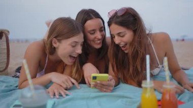 İki kız arkadaş plajda sosyal medyayı kontrol etmek için mobil bir cihazı paylaşıyorlar. Genç kadınlar akıllı telefonları kontrol eder ve çok gülerler. Telefonda video izlerken birlikte gülümserler..