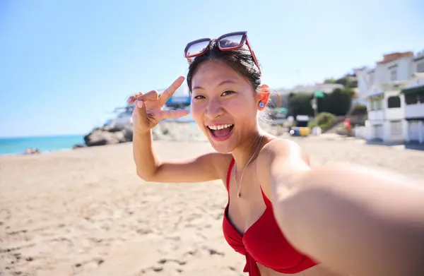 Young Asian Chinese woman in bikini smiling while taking a summer selfie on the beach. Vacation portrait of happy girl generation z female looking at camera cheerful makes peace gesture with hand