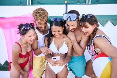 Bir grup çok ırklı mayo giymiş genç insan cep telefonlarını kullanarak gülüyorlar. Arkadaşlarımız yaz tatillerinde cep telefonlarında komik videolar izliyorlar.