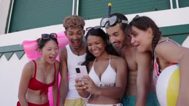 Bir grup çok ırklı mayo giymiş genç insan cep telefonlarını kullanarak gülüyorlar. Arkadaşlarımız yaz tatillerinde cep telefonlarında komik videolar izliyorlar.