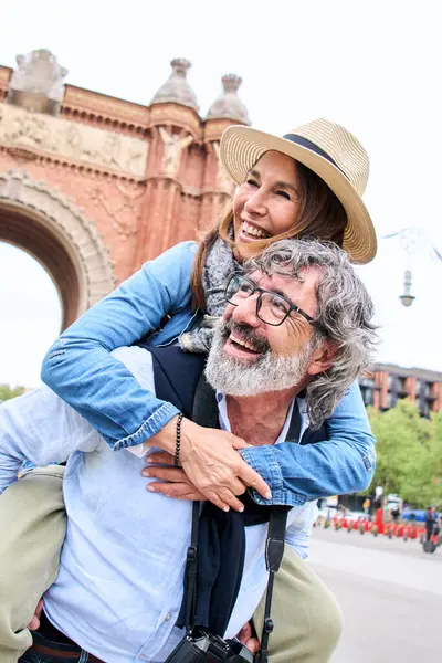 Vertical. Smiling senior couple in city street. Middle aged happy tourists enjoying weekend getaway together. Cheerful man with gray hair gives piggyback ride to woman in hat. Love relationship.