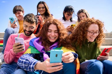 Gülümseyen genç arkadaşlar cep telefonlarını kullanarak eşcinsel onur festivalini kutluyorlar. Gökkuşağı bayraklı mutlu LGBT halkı dışarıda sosyal ağları izliyor ve kontrol ediyor