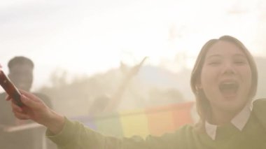 Heyecanlı genç bir Asyalı kız, günbatımında bir duman fişeğiyle kollarını kaldırıyor ve yanında özgürlük ve mutluluğu ifade eden çeşitli insanlar var. LGBT topluluk kutlamaları ve gay onur günü sembolleri