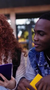 Üniversite binasının dışındaki merdivenlerde oturan üç genç uluslararası öğrencinin dikey videosu. Arkadaşlar kampüste dosyalar, çalışma kitapları ve notlarla mutlu bir şekilde bir araya geldiler.