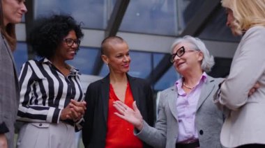 Düşük açı. Çeşitli yaş ve ırktan neşeli iş kadınlarından oluşan bir ekip ofis binasının dışında konuşarak ve sessizce iletişim kurarak mutlular. Resmi takım elbiseli profesyonel kadın girişimciler.