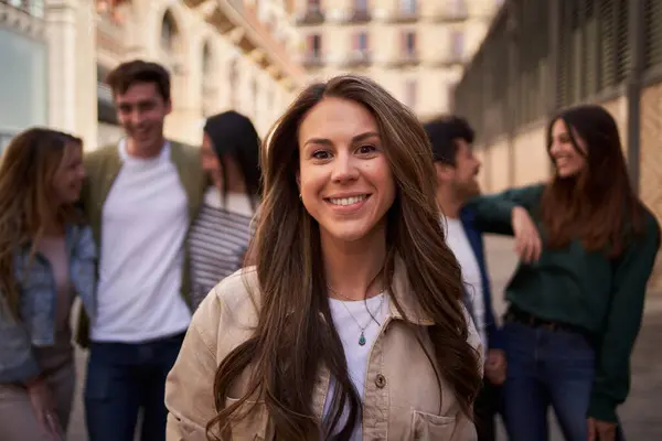 Portrait of Caucasian attractive woman looking smiling at camera in front unfocused group of people at meeting friendship. Young beautiful female posing happy outdoors with friends in background
