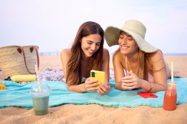 Havlunun üzerinde uzanmış, kumsalda eğlenen iki en iyi arkadaş. Birkaç genç bayan mayo giyip gülüyor. Ağ bağımlısı Z jenerasyonu insanları