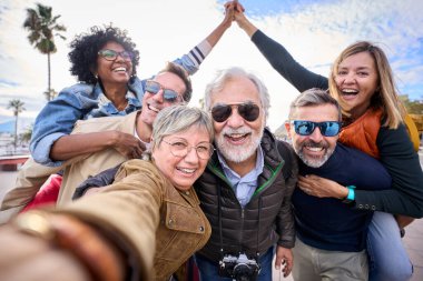 Group diverse funny mature tourist friends posing piggyback with hands joined in air taking photo selfie together with front camera on travel outdoor. Adult happy six excited people enjoy holidays  clipart