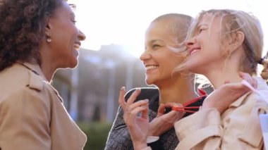 Yakın planda üç olgun kadın arkadaş bir araya gelip şehrin üzerinde gülüşerek sohbet ediyorlar. Çok ırklı kadın grup toplantısı güneşli açık havada kağıt alışveriş torbaları tutuyor. Pazarlık ve alışveriş düşkünleri