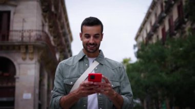 Avrupa 'nın Barselona şehrinde sokakta mutlu bir cep telefonu arayan genç beyaz bir adam yürüyor. Kırmızı hücre kullanarak gezen neşeli erkek turist açık hava seyahati deneyiminin tadını çıkarıyor