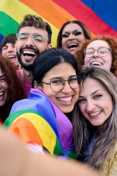 Dikey. Selfie LGBT grubu gençleri gey gurur gününü gökkuşağı bayrağını bir arada tutarak kutluyor. Homoseksüel toplum gülümsüyor ve neşeli bir oto portre çekiyor. Lezbiyen çift ve arkadaş nesli z