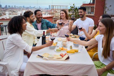 Kırmızı şarap içen ve çatıda yemek yiyen bir grup mutlu genç. Binlerce yıllık arkadaşlar birlikte dışarıda alkollü içecekler içip tatillerde öğle yemeğinin tadını çıkarıyorlar.