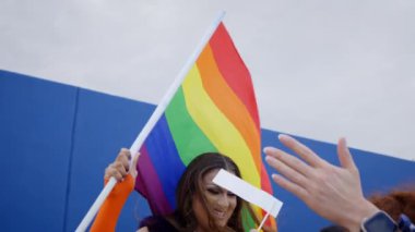 Mavi bir duvarın önünde neşeli bir bayrağın bayrağını sallayan genç, gülümseyen bir travesti. LGBT özgür aşk ve seks festivalini kutlayan mutlu transseksüeller topluluğu