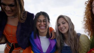 Düşük açı. Bir grup mutlu genç, gökkuşağı bayraklarıyla kucaklaşan kameraya bakıp gülüyor ve dışarıda LGBT etkinliğinin eğlenceli anlarını paylaşıyor.