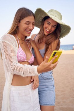 Sahilde telefonda eğlenen iki güzel arkadaş. Birlikte genç ve mutlu kadınlar mayo giyip gülüyorlar. Sosyal ağlara bağımlı birkaç çekici kız nesli.