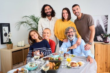 Beyaz bir ailenin gülümseyen bir portresi. Kapalı alanda kucaklaşan kameralara bakıyor. Nesiller boyu neşeli insanlar, evde birlikte yemek yemek için masanın etrafında toplandılar.