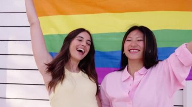 Açık havada gökkuşağı bayrağı taşıyan aşık genç çift lezbiyen kızlar. İki tane gülen, çok ırklı, tatlı kadın beyaz geçmişli, gurur dolu bir kutlama yapıyorlar. Kadın eşcinseller arasındaki arkadaşlık