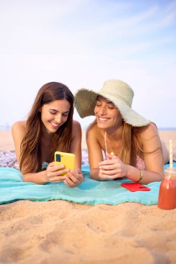 İki en iyi arkadaşın havlunun üzerinde yatıp eğlenirken çekilmiş dikey bir fotoğrafı. Birkaç genç bayan mayo giyip gülüyor. Ağ bağımlısı Z jenerasyonu insanları