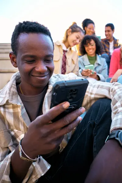 Vertical. Smiling black guy using smart phone. African American man holding cell outdoor with friends background. Carefree young people having fun together. Technology addicted generation