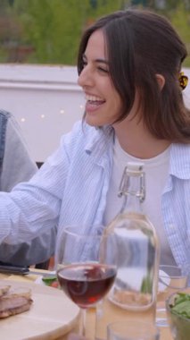 Dikey. Çeşitli genç arkadaşlar terasta bir yemekte kırmızı şarapla kadeh kaldırarak kutluyorlar. İnsanlar gülerek gülerek ilkbahar günü dışarıda toplandılar. Yemek, içecek ve çatıda eğlenmek.