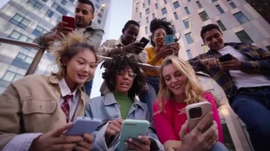 Group of young people watching something at phone sitting on stairs. Smiling multicultural friends looking social media outside the university campus. Students in their free time.