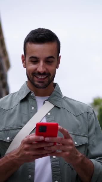 ヨーロッパのバルセロナの街の通りで幸せな携帯電話を見て歩いている垂直若い白人魅力的な男性 赤いセルを使用して散歩する楽しい男性観光客は 屋外旅行の観光をタイプを楽しんでいます — ストック動画