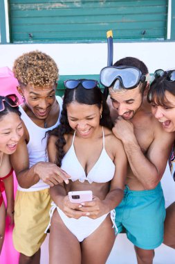Mayolu genç ve çok ırklı insanlardan oluşan dikey fotoğraf grubu gülüyor ve cep telefonuna bakıyorlar. Arkadaşlarımız yaz tatillerinde cep telefonlarında komik videolar izliyorlar.